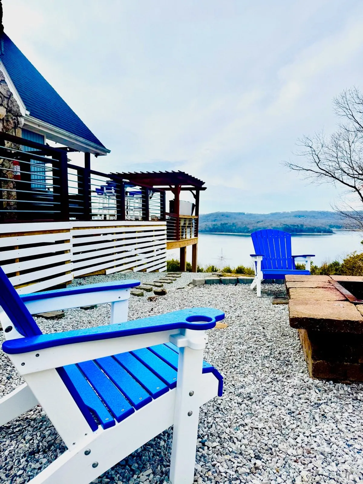A cozy firepit area is framed by blue Adirondack chairs, set just steps from the lake. The background reveals peaceful water and sky, suggesting an ideal spot for stargazing, sharing stories, or enjoying s'mores by the fire.