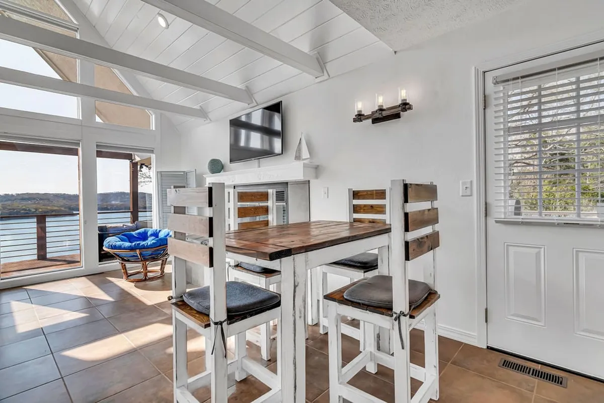 A bright, open dining area with a wooden dining table and high-backed chairs sits in front of expansive windows that frame a shimmering lake view. The decor is minimalist and clean, and there's a television in the background for added comfort and entertainment. Natural light floods the space, creating a warm and welcoming ambiance.