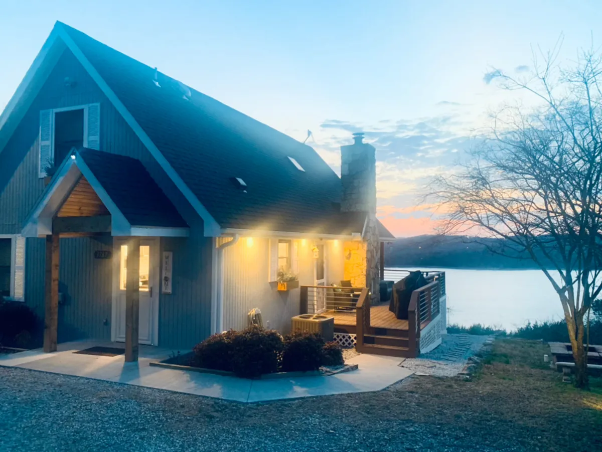 The charming exterior of Blue Moon Cottage is shown, highlighting its wrap-around porch with inviting seating and expansive water views in the background. The scene radiates coziness, with a hint of an evening gathering spot by the fire pit.