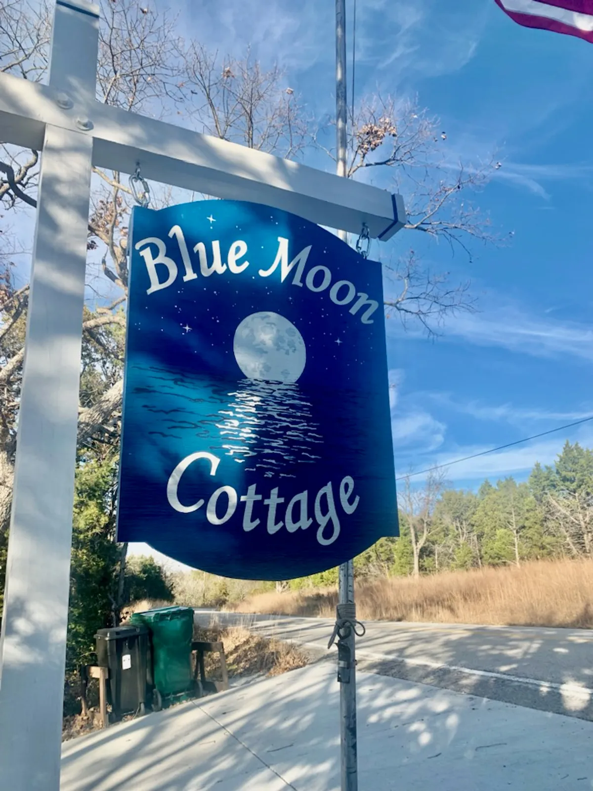 This image features a clear, welcoming sign with 'Blue Moon Cottage' set against a backdrop of a clear blue sky and the moon overhead. The surrounding area appears tranquil and lightly wooded, suggesting a private and natural setting.