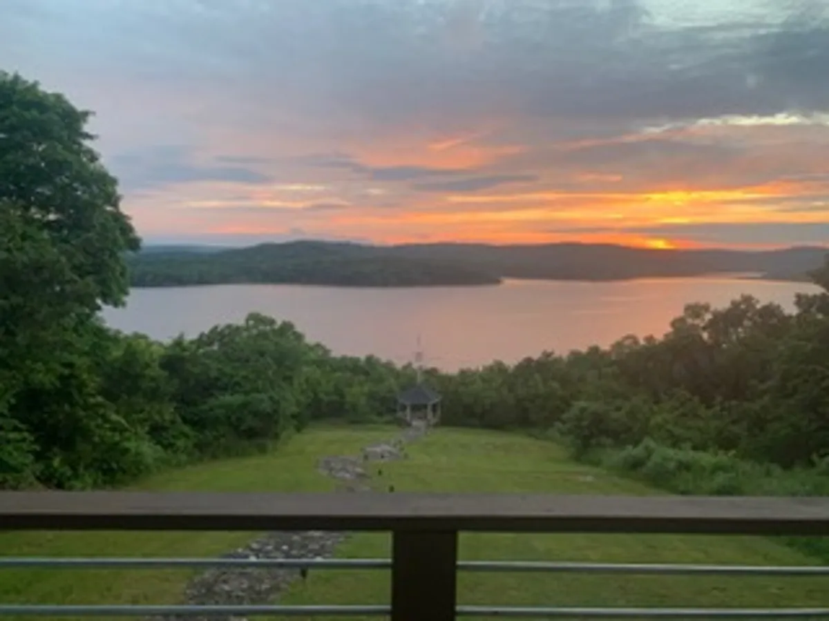 A peaceful lake scene with a gazebo perched by the water's edge. The sun is setting, coloring the sky in soft hues. Trees surround the area, and the fire pit is visible for evening gatherings, offering a serene view and opportunities for nature exploration.