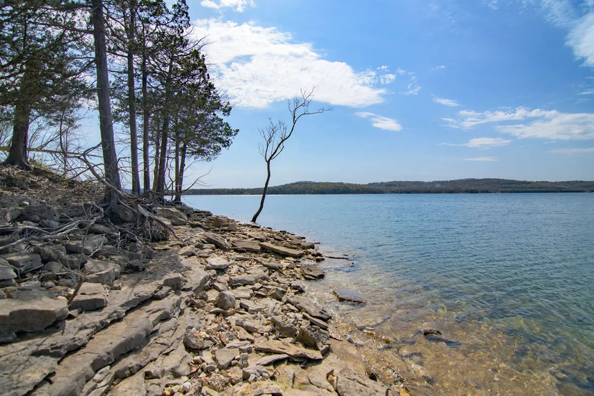 A picturesque view of the lake, rocky shoreline, scattered trees, and distant hills, ideal for fishing, picnicking, hiking, and stargazing. The tranquil mood reflects seasonal outdoor exploration and the beauty of natural surroundings.