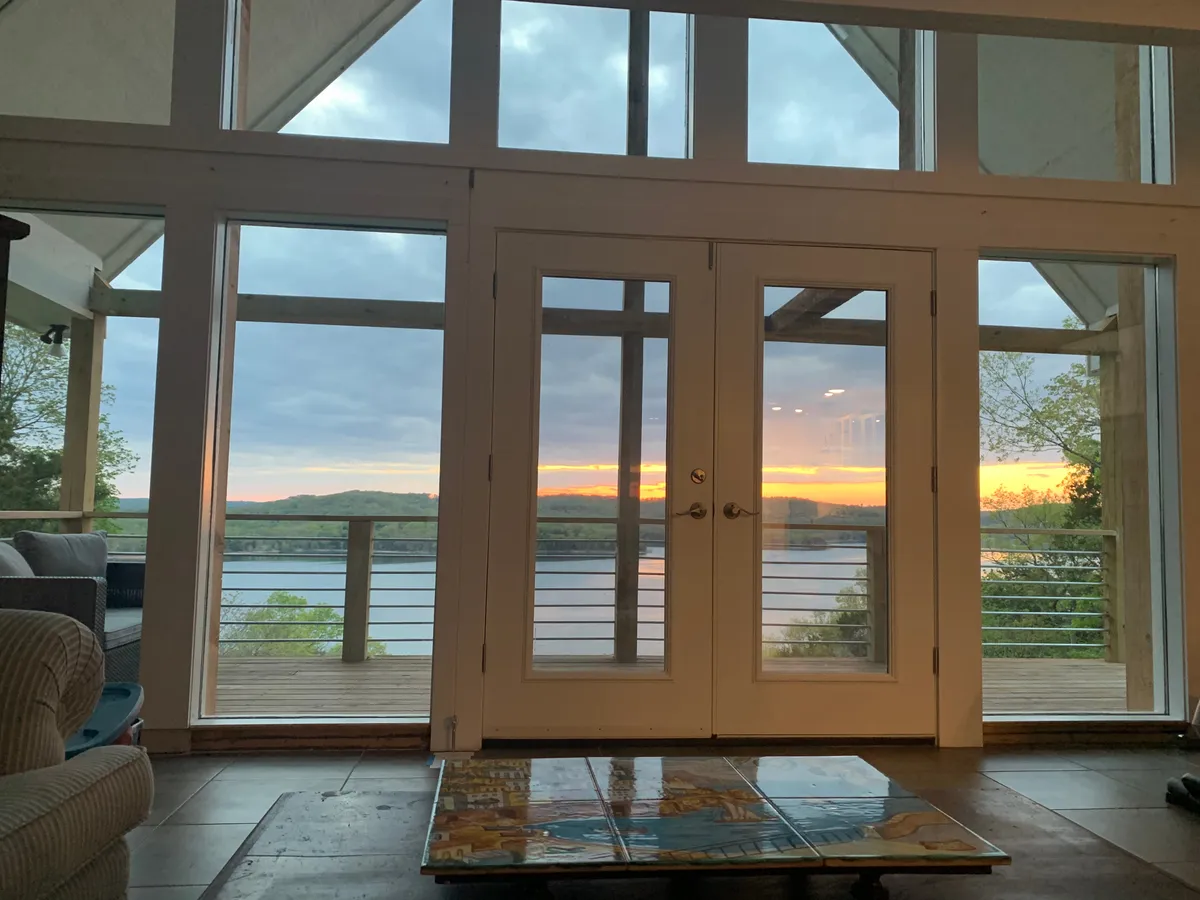 An inviting family room featuring a reclining sofa positioned to take in the golden glow of a sunset through large glass doors. The room offers effortless access to the outdoors, seamlessly connecting the interior with the wrap-around porch and water views beyond.