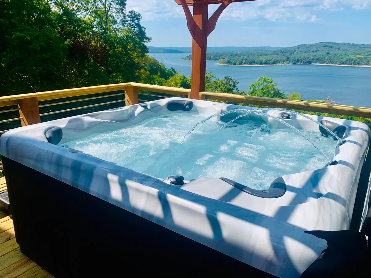 A bubbling hot tub sits on a deck with panoramic views of the lake and is surrounded by lush greenery for privacy. The setting feels intimate and serene, ideal for enjoying a soak while watching the sky change color at sunset.