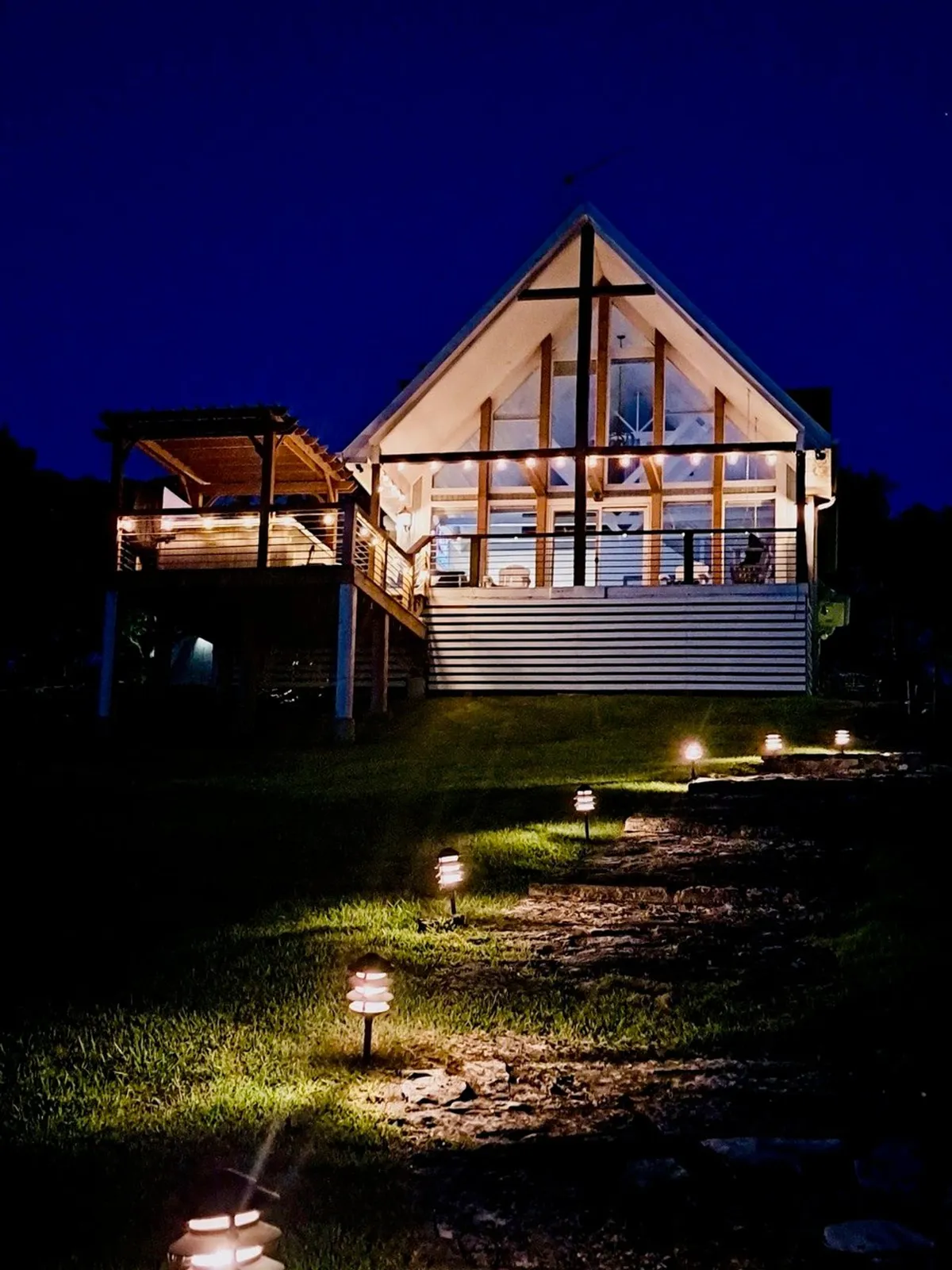 Blue Moon Cottage glows warmly in the evening, with pathway lights guiding guests to the inviting deck area. The illuminated house is surrounded by trees, and the deck hints at space for gatherings around a fire pit or enjoying the sunset.