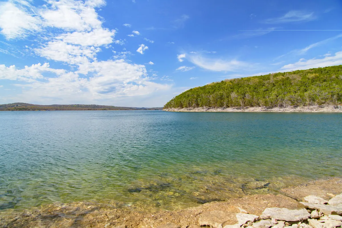 A tranquil scene of calm Table Rock Lake bordered by a green hillside and rocky shoreline under a blue sky with clouds. The setting evokes opportunities for water activities like fishing, boating, and hiking along the shore, while offering glimpses of local wildlife and vibrant seasonal change.