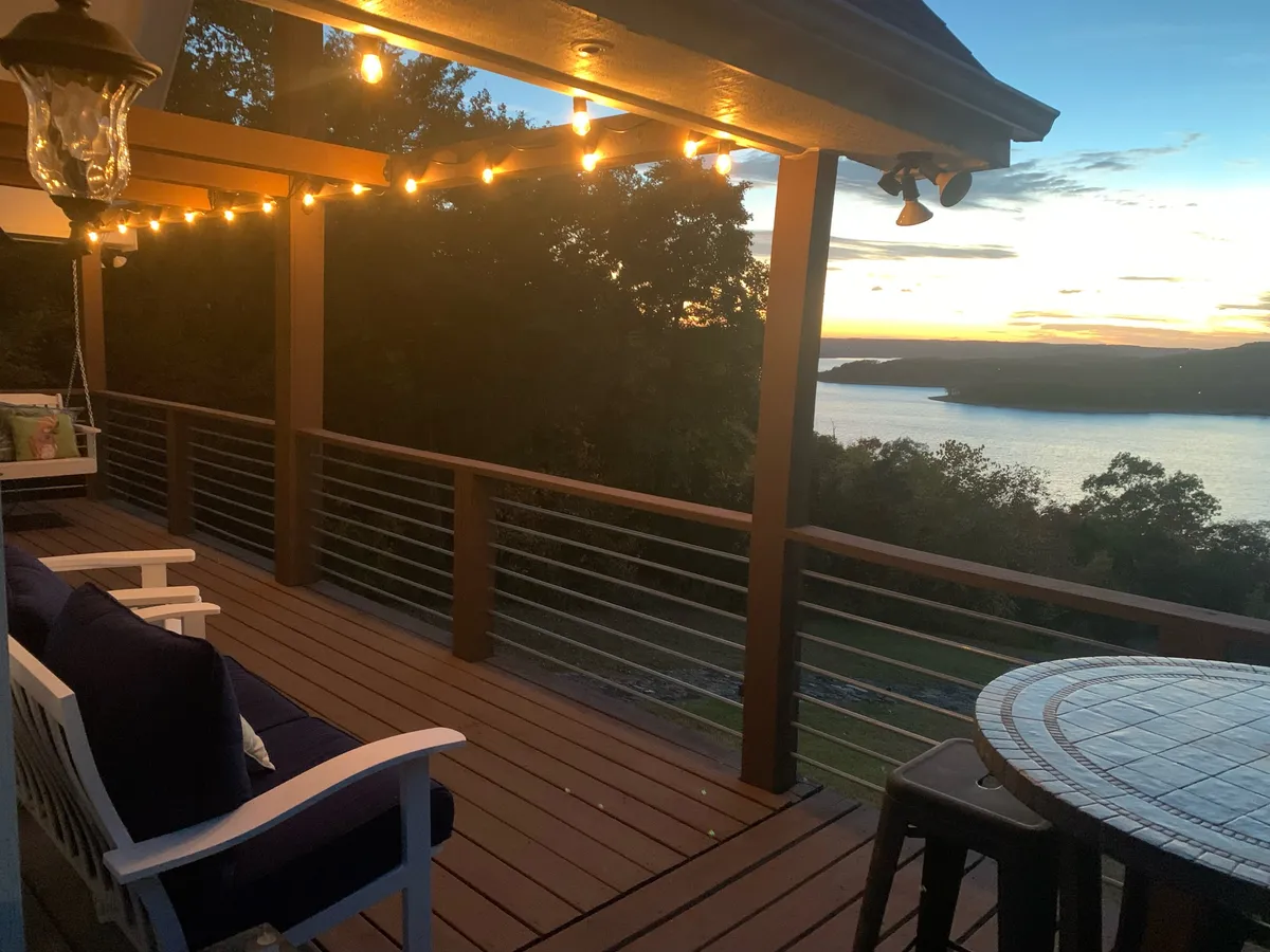 A wide wooden deck wraps around the cottage, strung with glowing lights, and overlooks a tranquil lake. Outdoor furniture is arranged for both dining and lounging, offering a comfortable space to soak in sunset views or gather for celebrations.