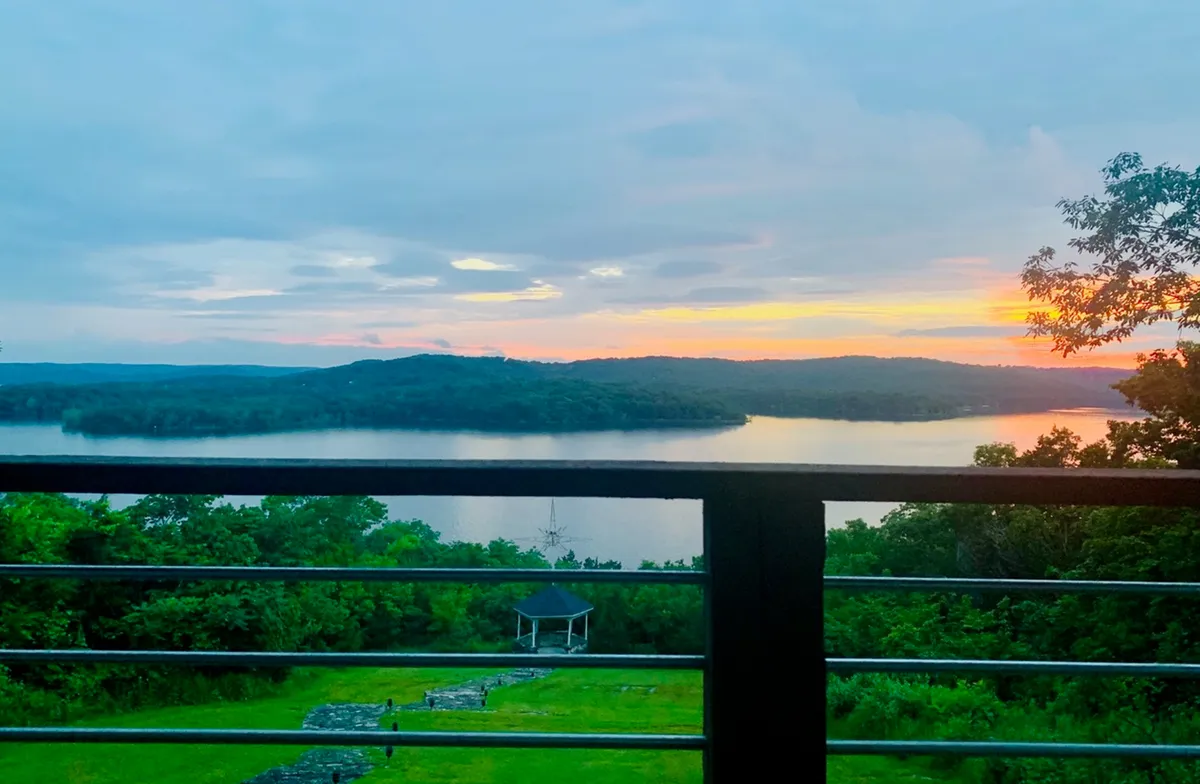 A breathtaking sunset spills vibrant colors across the sky above the lake, with a gazebo nestled among lush greenery in the foreground. The peaceful setting is perfect for quiet reflection or sharing a romantic moment while the sun dips below the horizon.