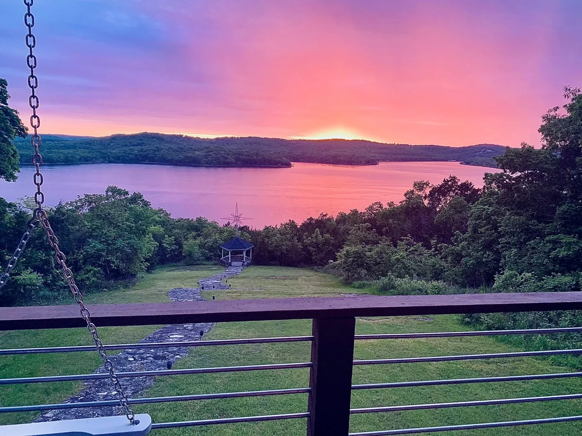 A scenic view of a sunset over the lake, framed by lush greenery and a stone pathway leading toward a gazebo in the distance. The atmosphere is peaceful and beckons exploration or quiet reflection in nature.