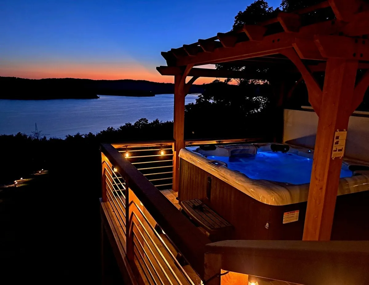 The image features the cottage's outdoor hot tub, perfectly positioned to overlook a stunning panoramic lake view at sunset. The warm glow of the sunset reflects off the water, creating a serene atmosphere for relaxation and stargazing. The cottage exterior is inviting, and the setting feels private and intimate, ideal for unwinding or sharing memorable moments with loved ones.