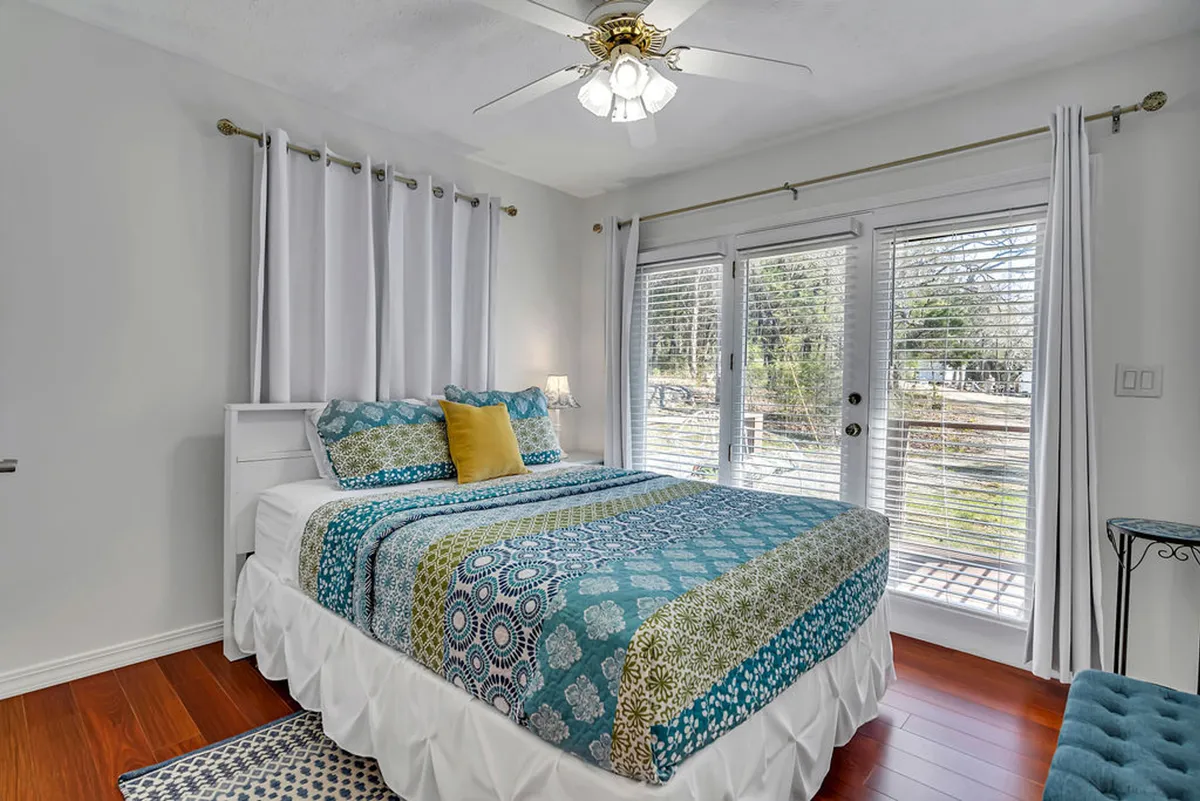 A cozy bedroom with a bed featuring colorful bedding is bathed in natural light from large windows. A decorative side table holds a cup of coffee, suggesting a peaceful start to the day.